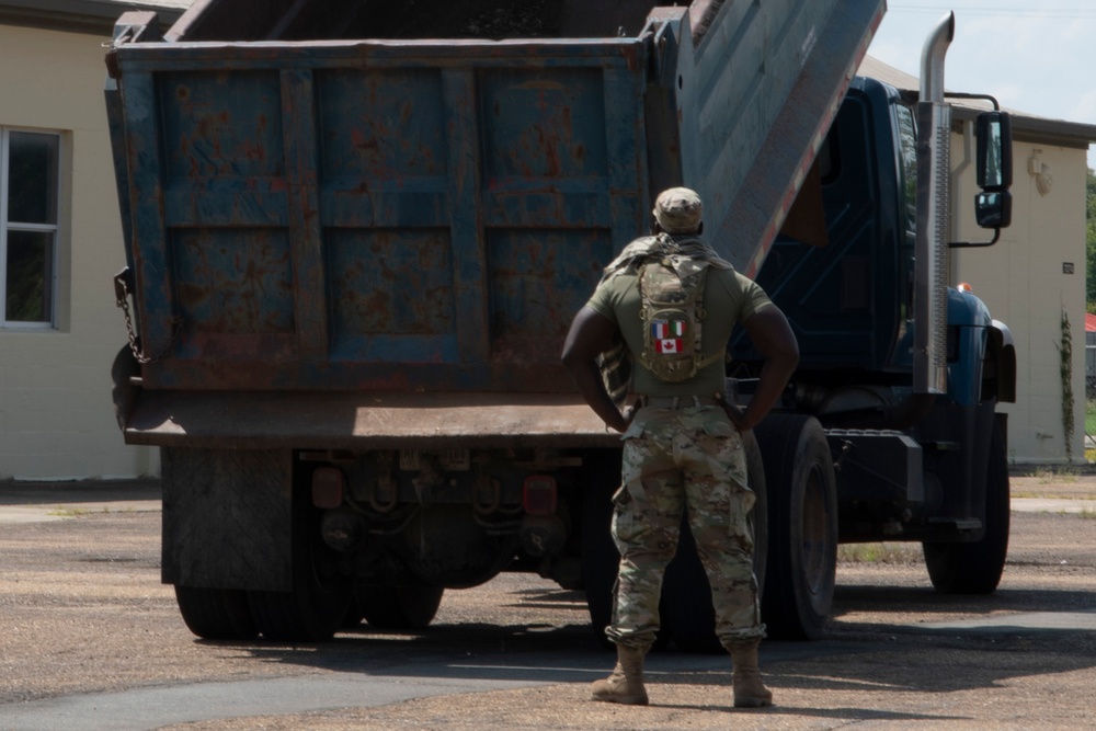 A military personnel member in camouflage uniform stands facing a large, weathered blue dump truck with its bed raised. The soldier is wearing a backpack displaying what appear to be French and Canadian flag patches. The scene appears to take place in an outdoor area near a building, suggesting a logistics or checkpoint operation.