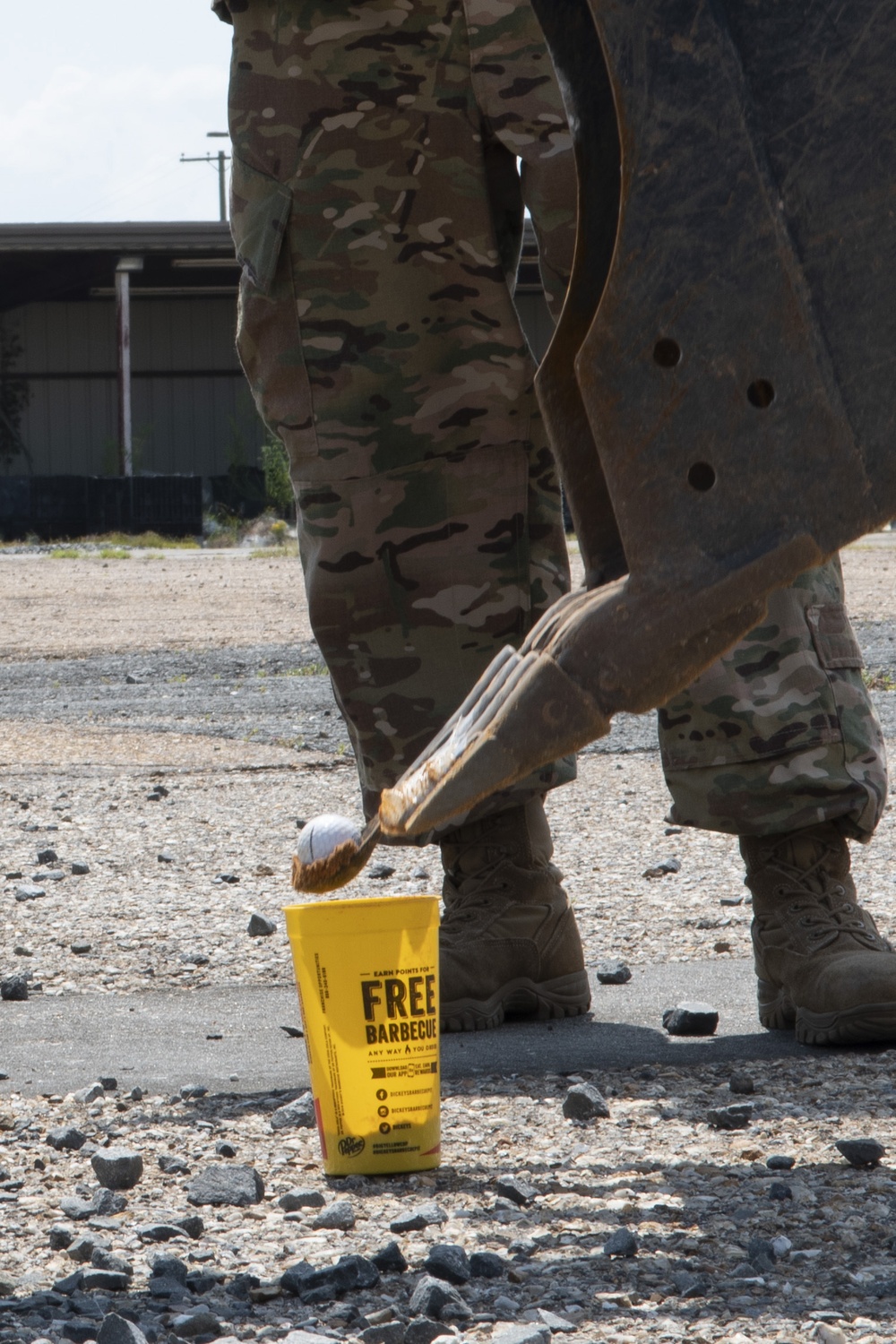 A person wearing military camouflage (OCP/MultiCam pattern) uniform and tan combat boots is standing on a gravel/asphalt surface. A yellow cup branded with 'FREE BARBECUE' text (appearing to be from Dickey's Barbecue) is on the ground near their feet. A large metal implement, possibly an excavator bucket or heavy equipment attachment, is visible beside the person.