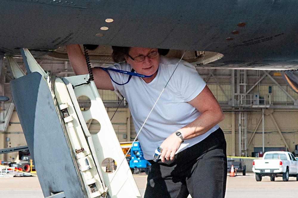 A technician or maintenance worker is inspecting or servicing the underside of a large aircraft on an airfield tarmac. The person is crouched beneath the fuselage, holding a tool and wearing a blue lanyard with an ID badge. A military or commercial aviation hangar is visible in the background.