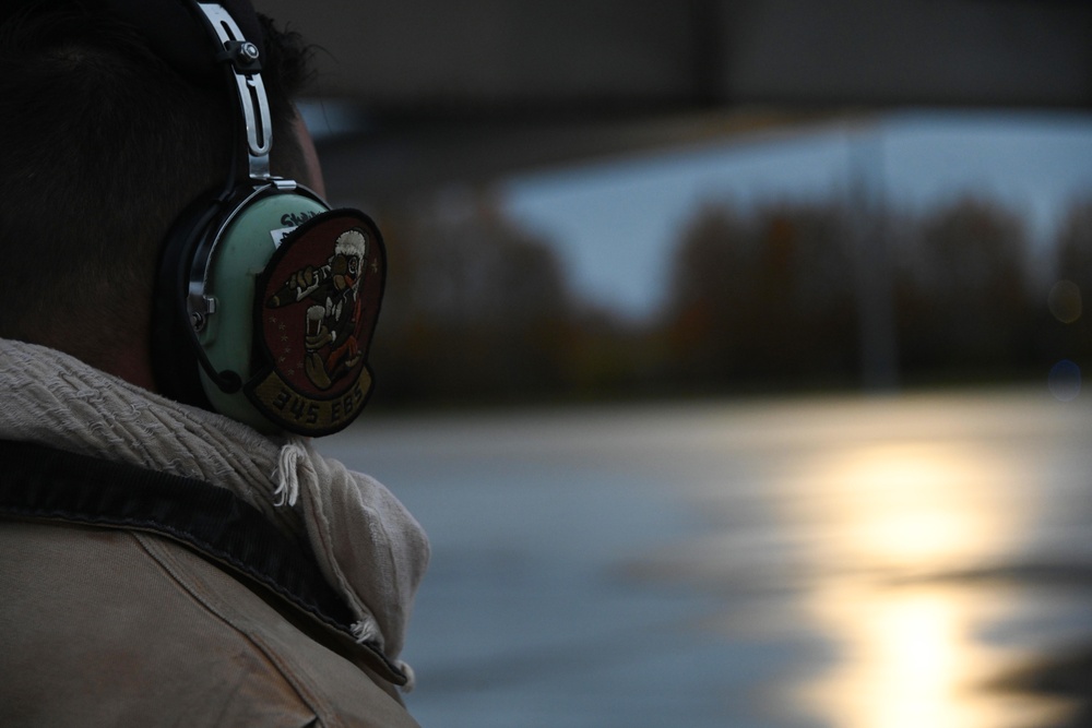 A person wearing military-style hearing protection/headset is photographed from behind, facing a wet tarmac or airfield surface. The headset features a unit patch with a cartoon mascot figure and text reading '345 EES' on a banner. The background shows a blurred airfield environment with trees and reflected light on the wet surface.