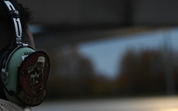 A person wearing military-style hearing protection/headset is photographed from behind, facing a wet tarmac or airfield surface. The headset features a unit patch with a cartoon mascot figure and text reading '345 EES' on a banner. The background shows a blurred airfield environment with trees and reflected light on the wet surface.