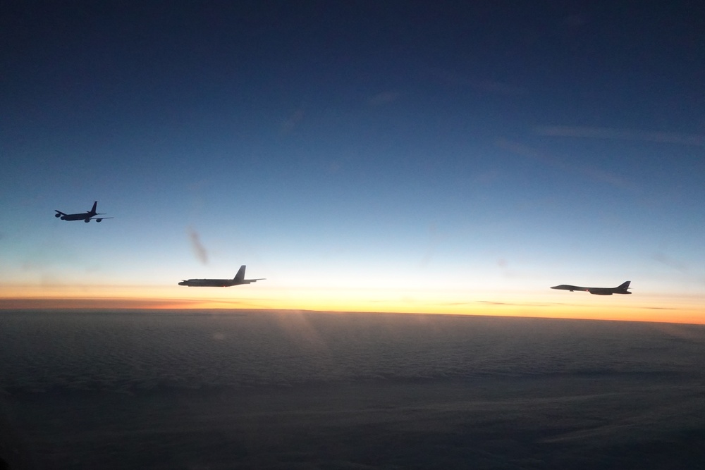 Three military aircraft are flying in loose formation above a cloud layer during twilight or dawn conditions. The aircraft are captured as silhouettes against a gradient sky transitioning from deep blue at the top to orange and yellow near the horizon. The scene appears to be taken from a fourth aircraft, suggesting a multi-aircraft mission or formation flight.