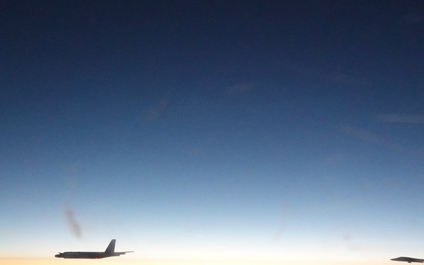 Three military aircraft are flying in loose formation above a cloud layer during twilight or dawn conditions. The aircraft are captured as silhouettes against a gradient sky transitioning from deep blue at the top to orange and yellow near the horizon. The scene appears to be taken from a fourth aircraft, suggesting a multi-aircraft mission or formation flight.