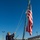 A uniformed sailor wearing a face mask and tan cap is raising the American flag on what appears to be the deck of a U.S. Navy vessel moored at a pier. The flag is fully unfurled and flying against a clear, deep blue sky. A second person is partially visible behind the primary subject, and port infrastructure is visible in the background.