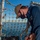 A U.S. Navy sailor in blue working uniform is performing maintenance or rigging work on the deck of a naval vessel. The sailor is wearing a black face mask, a tan cap with 'DDG' visible, and has a rate insignia patch on the uniform. The work involves handling wire rope/cable and hardware fittings near a safety net on the ship's deck, with open water visible in the background.