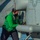 A military aviation technician or deck crew member wearing a green jersey and black face mask is performing maintenance or inspection work on a naval helicopter. The individual is working near the fuselage of what appears to be a U.S. Navy helicopter, visible by partial 'NAV' lettering on the aircraft. A cylindrical pod or external store is mounted beneath the aircraft's fuselage.