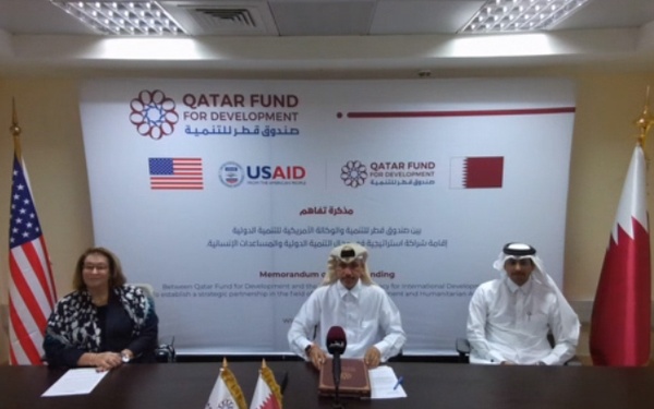 A virtual signing ceremony of a Memorandum of Understanding (MOU) between the Qatar Fund for Development and USAID, establishing a strategic partnership in international development and humanitarian assistance. The image shows two simultaneous video feeds: the Qatar side (Hub-01, 10th floor DG's Conference Room) with three officials seated at a table, and the USAID side featuring Max Primorac holding up the signed document. Both organizations' logos, flags, and branding are prominently displayed.