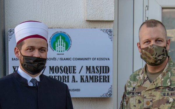 Two individuals stand in front of a placard identifying the Islamic Community of Kosova / Bashkësia Islame e Kosovës, specifically a mosque named Masjid Bedri A. Kamberi. One person is wearing Islamic clerical attire with a white turban and black robe, while the other is in U.S. Army multicam uniform. Both are wearing face masks, suggesting this photo was taken during the COVID-19 pandemic period.