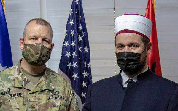 Two individuals pose together in front of flags, including the United States flag and what appears to be the Albanian flag. One person is wearing a U.S. Army uniform with the name tape 'VAN SLOTEN' and a chaplain's cross insignia, while the other is dressed in traditional Islamic clerical attire including a white turban and dark robe. Both individuals are wearing black face masks.