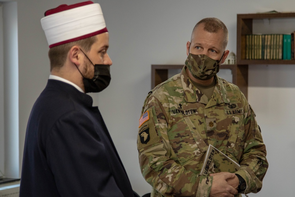 A U.S. Army chaplain in OCP camouflage uniform is engaged in conversation with a Muslim cleric wearing traditional white and red religious headwear (takke/kalpak). Both individuals are wearing face masks, suggesting this meeting took place during the COVID-19 pandemic period. The setting appears to be an office or religious facility with bookshelves visible in the background.