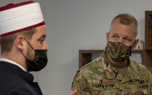 A U.S. Army chaplain in OCP camouflage uniform is engaged in conversation with a Muslim cleric wearing traditional white and red religious headwear (takke/kalpak). Both individuals are wearing face masks, suggesting this meeting took place during the COVID-19 pandemic period. The setting appears to be an office or religious facility with bookshelves visible in the background.