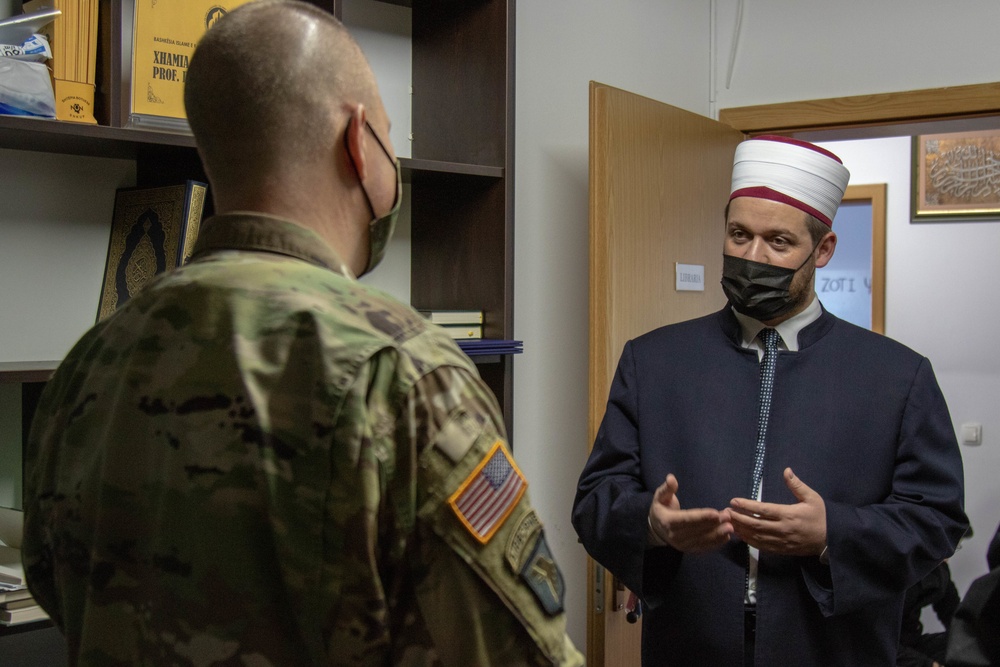 A U.S. Army soldier in camouflage uniform is meeting with a Muslim cleric or imam wearing traditional religious attire including a white and red cylindrical hat (imamah/sarık). Both individuals are wearing black face masks, suggesting the meeting took place during the COVID-19 pandemic. The setting appears to be an office or library within a mosque or Islamic institution.