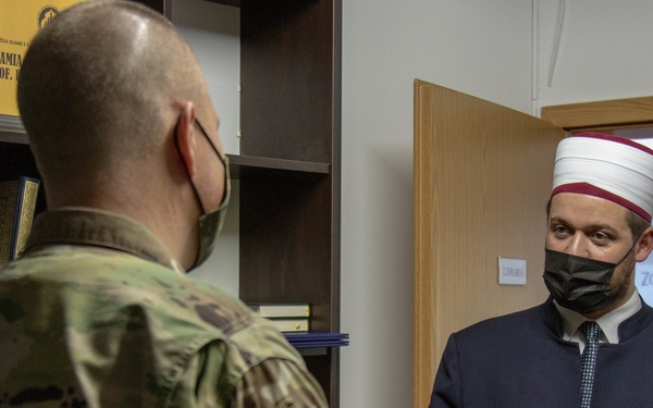 A U.S. Army soldier in camouflage uniform is meeting with a Muslim cleric or imam wearing traditional religious attire including a white and red cylindrical hat (imamah/sarık). Both individuals are wearing black face masks, suggesting the meeting took place during the COVID-19 pandemic. The setting appears to be an office or library within a mosque or Islamic institution.