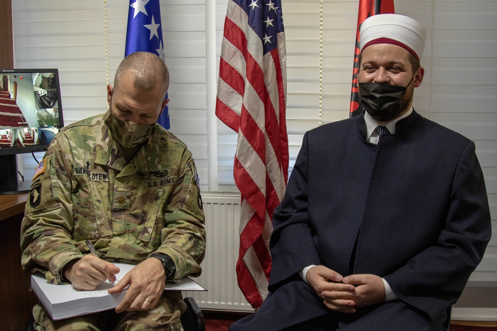 Two individuals are seated side by side in what appears to be an official meeting room. A U.S. Army soldier in camouflage uniform is writing on a document, while a religious leader wearing traditional Islamic clerical attire and a white turban looks toward the camera. Both individuals are wearing black face masks, and multiple national flags are visible in the background.