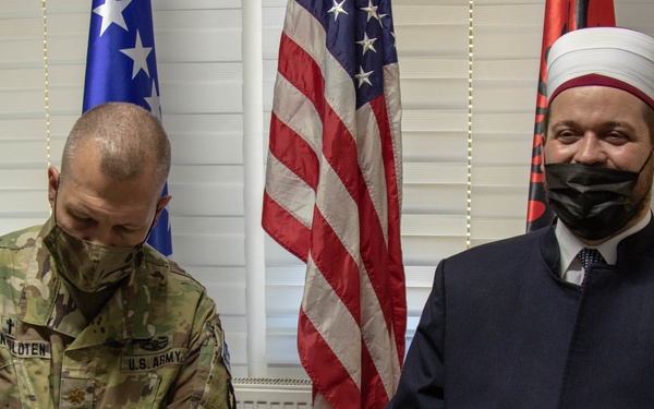 Two individuals are seated side by side in what appears to be an official meeting room. A U.S. Army soldier in camouflage uniform is writing on a document, while a religious leader wearing traditional Islamic clerical attire and a white turban looks toward the camera. Both individuals are wearing black face masks, and multiple national flags are visible in the background.