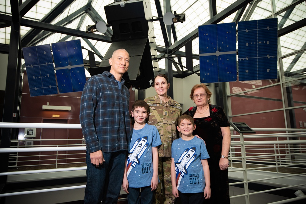 Chief Master Sergeant April Brittain poses with family in front of satellite static model after transfer ceremony into USSF