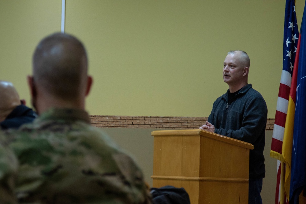 A civilian man in a dark fleece jacket stands at a wooden podium, appearing to address an audience that includes at least one uniformed military personnel in camouflage. The setting appears to be an official military briefing or ceremony room, indicated by the presence of the U.S. flag and what appears to be a NATO or unit flag in the background. The foreground shows the back of a soldier in MultiCam/OCP uniform listening to the speaker.