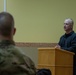 A civilian man in a dark fleece jacket stands at a wooden podium, appearing to address an audience that includes at least one uniformed military personnel in camouflage. The setting appears to be an official military briefing or ceremony room, indicated by the presence of the U.S. flag and what appears to be a NATO or unit flag in the background. The foreground shows the back of a soldier in MultiCam/OCP uniform listening to the speaker.