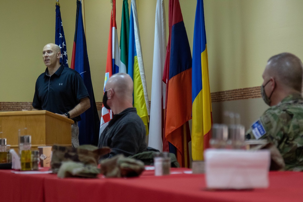 A man in a black polo shirt speaks at a wooden podium during what appears to be a multinational military or diplomatic briefing. Multiple international flags are displayed in the background, and attendees including at least one uniformed military personnel wearing a face mask are seated at a red-clothed table in the foreground. The setting suggests a formal conference or meeting room.
