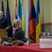 A man in a black polo shirt speaks at a wooden podium during what appears to be a multinational military or diplomatic briefing. Multiple international flags are displayed in the background, and attendees including at least one uniformed military personnel wearing a face mask are seated at a red-clothed table in the foreground. The setting suggests a formal conference or meeting room.