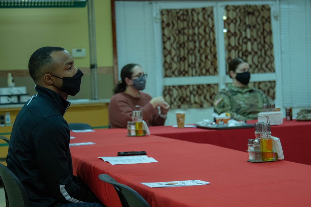 Three individuals are seated at a red-tablecloth-covered table in what appears to be a dining facility or meeting room. All three are wearing face masks, suggesting this was taken during the COVID-19 pandemic period. One person in the background is wearing military camouflage uniform, while the other two are in civilian attire.