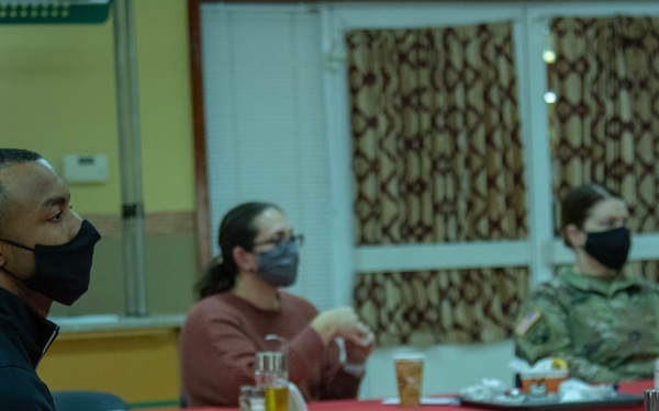 Three individuals are seated at a red-tablecloth-covered table in what appears to be a dining facility or meeting room. All three are wearing face masks, suggesting this was taken during the COVID-19 pandemic period. One person in the background is wearing military camouflage uniform, while the other two are in civilian attire.