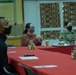 Three individuals are seated at a red-tablecloth-covered table in what appears to be a dining facility or meeting room. All three are wearing face masks, suggesting this was taken during the COVID-19 pandemic period. One person in the background is wearing military camouflage uniform, while the other two are in civilian attire.