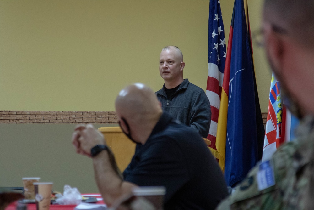 A man in a gray zip-up pullover appears to be speaking or presenting at a formal meeting or briefing. Several other individuals are seated in the foreground, with one wearing a face mask. Multiple national and organizational flags are displayed in the background, including the United States flag, German flag, NATO flag, and what appears to be a Croatian flag.