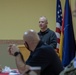 A man in a gray zip-up pullover appears to be speaking or presenting at a formal meeting or briefing. Several other individuals are seated in the foreground, with one wearing a face mask. Multiple national and organizational flags are displayed in the background, including the United States flag, German flag, NATO flag, and what appears to be a Croatian flag.