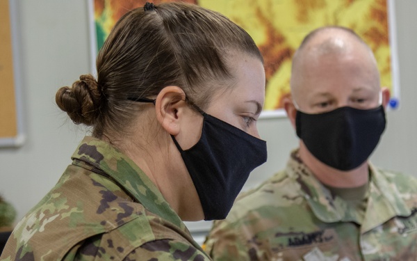 Two U.S. Army soldiers wearing black face masks and OCP (Operational Camouflage Pattern) uniforms are reviewing a document together in what appears to be an office or administrative setting. The female soldier in the foreground displays a KFOR Headquarters GEO patch and a U.S. flag patch on her sleeve, indicating assignment to Kosovo Force. Both soldiers appear to be engaged in a briefing or administrative review task.