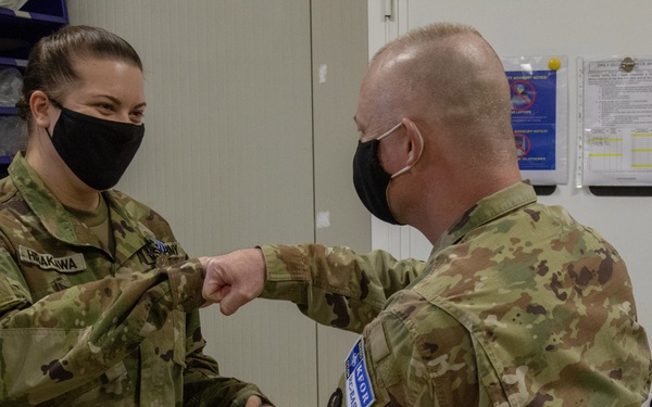 Two military personnel in OCP (Operational Camouflage Pattern) uniforms are performing an elbow bump greeting, a COVID-era alternative to handshakes. Both individuals are wearing black face masks, and one is holding a green folder. The setting appears to be an indoor military facility or office space.
