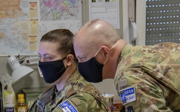 Two military personnel in U.S. Army OCP (Operational Camouflage Pattern) uniforms are working together at a desk in what appears to be a military office or operations room. Both individuals are wearing black face masks and display KFOR (Kosovo Force) and RC-EAST patches on their uniforms. They appear to be reviewing documents or working at a workstation together.