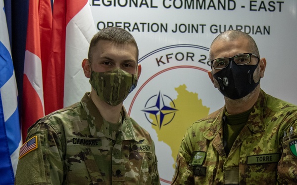 Two military personnel, one from the U.S. Army and one from the Italian Army, are posing together while holding a NATO challenge coin. The ceremony takes place in front of a backdrop reading 'Regional Command - East, Operation Joint Guardian, KFOR 28' with the NATO compass rose logo. Both individuals are wearing face masks and are surrounded by multiple national flags.