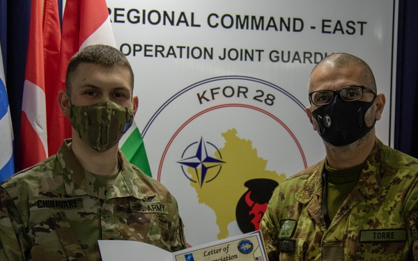 Two military personnel are posing together holding a 'Letter of Appreciation' in front of a KFOR 28 Regional Command East / Operation Joint Guardian backdrop. The soldier on the left wears a US Army uniform with the name tape 'CHINAVARE', while the soldier on the right wears an Italian Army uniform with the name tape 'TORRE'. Both are wearing black face masks, suggesting this photo was taken during the COVID-19 pandemic period.