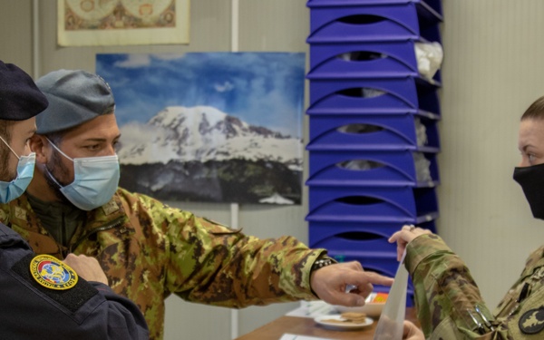 Three military/law enforcement personnel wearing face masks are engaged in a document exchange at an indoor desk or counter. Two individuals in camouflage uniforms and one in a dark navy uniform are interacting in what appears to be an administrative or coordination setting. Visible patches suggest NATO KFOR (Kosovo Force) affiliation on at least one uniform.