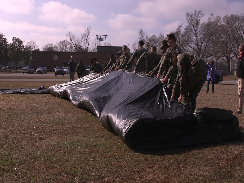 A group of military personnel in camouflage uniforms are working together to fold or deploy a large black tarpaulin or cover on an open grassy field. In the background, a tiltrotor aircraft (consistent with a V-22 Osprey) is visible in flight. The scene appears to take place on a military installation, with civilian vehicles and buildings visible in the background.
