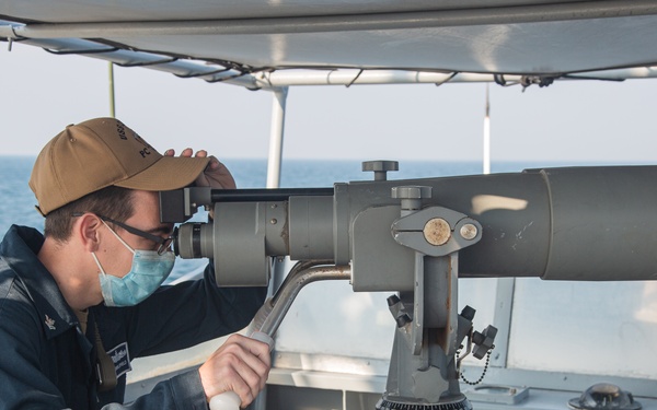 A naval personnel member is using a large mounted binocular/telescope device on the bridge wing or observation deck of a military vessel. The individual is wearing a face mask, glasses, a tan/khaki cap, and a dark navy uniform. The scene takes place at sea with calm blue water visible in the background.