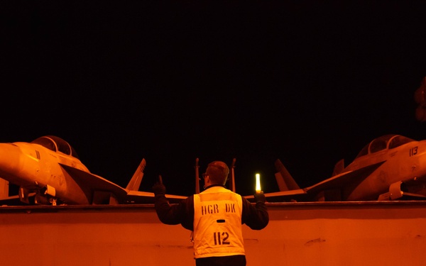 A night scene on what appears to be an aircraft carrier flight deck, illuminated by orange/red lighting. A deck crew member wearing a reflective vest marked 'HGR DK 112' is directing aircraft using illuminated wands. Two military jet aircraft, visible with markings '113' on one, are parked on the deck.