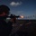A person wearing protective eyewear is firing an M16/M4-style rifle on what appears to be the deck of a naval vessel at dusk or dawn. A muzzle flash is visible at the end of the barrel, and a row of illuminated silhouette targets is visible downrange. This appears to be a live-fire qualification or training exercise conducted aboard a ship.