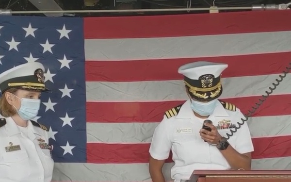 Two U.S. Navy officers in white dress uniforms stand at a podium aboard the USS Tortuga (LSD 46), a dock landing ship. Both officers are wearing face masks, suggesting this event took place during the COVID-19 pandemic. The setting appears to be an official ceremony, likely a change of command or similar formal naval event.