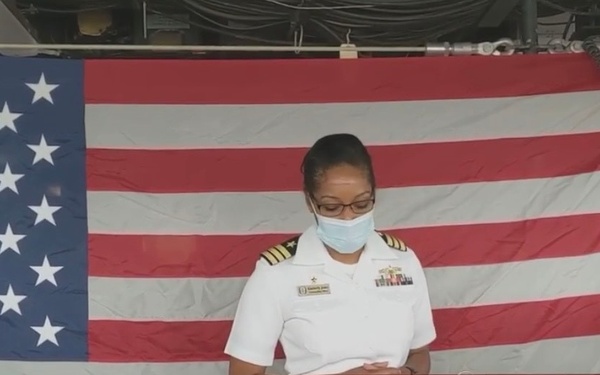 A U.S. Navy officer in white dress uniform stands at a podium aboard the USS Tortuga (LSD 46), a dock landing ship. The officer is wearing a face mask, suggesting this event took place during the COVID-19 pandemic period. A large American flag is displayed prominently in the background.