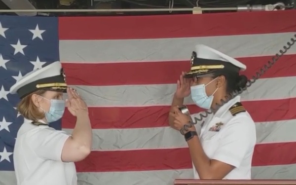 Two U.S. Navy officers in white dress uniforms are exchanging salutes at a podium bearing the name 'USS Tortuga (LSD 46)', a dock landing ship. Both individuals are wearing face masks, suggesting this ceremony took place during the COVID-19 pandemic. An American flag is displayed prominently in the background.
