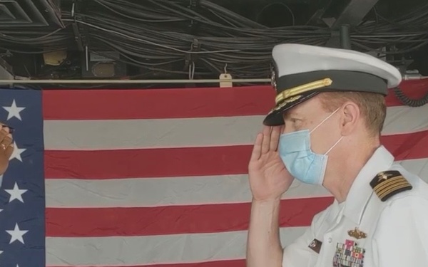 Two U.S. Navy officers in white dress uniforms are exchanging salutes aboard a naval vessel, with an American flag displayed in the background. A wooden podium bearing the partial name 'USS T...(L...)' is visible between them, suggesting a formal military ceremony. Both officers are wearing blue face masks, indicating this event took place during the COVID-19 pandemic era.