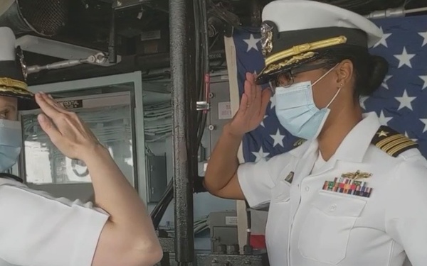 Two female U.S. Navy officers in white dress uniforms are exchanging salutes aboard what appears to be a naval vessel. Both are wearing blue face masks, consistent with COVID-19 era protocols. An American flag is displayed in the background, suggesting a formal military ceremony or change of command.