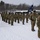 A formation of U.S. Army soldiers stands at attention in snowy winter conditions, wearing OCP (Operational Camouflage Pattern) uniforms and cold-weather gear including black balaclavas and gloves. A guidon bearer holds a blue unit flag marked 'HHC 3,' indicating Headquarters and Headquarters Company of a 3rd unit. The formation appears to be part of a formal military ceremony or muster at a snow-covered installation.