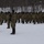 A large formation of military personnel in camouflage uniforms stands at attention or saluting in a snow-covered outdoor area. The formation is arranged in multiple rows facing a central figure, with the American flag and what appears to be a unit guidon visible on the left side. The setting appears to be a winter military ceremony or formation, likely at a training installation.