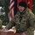 A U.S. Army soldier named Wignall is speaking at an outdoor podium during what appears to be a military ceremony in winter conditions. He is wearing MultiCam/OCP camouflage uniform with a black watch cap and displays a 'Mountain' shoulder sleeve insignia. An American flag and a red military unit guidon are visible in the background.