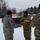 Two U.S. Army soldiers are conducting a military colors ceremony outdoors in snowy winter conditions. One soldier in UCP/ACU pattern camouflage and the other in OCP/Multicam pattern are handling a folded blue guidon or unit flag with gold fringe. A third soldier in OCP uniform is partially visible on the right side of the frame.