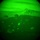 A night vision (NVG) captured image showing multiple parachutes deployed over a mountainous terrain. The circular green-tinted field of view is characteristic of image intensifier night vision optics. Numerous parachutes are visible at various altitudes, suggesting a mass tactical airborne operation.