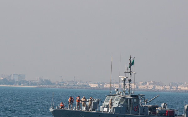 A gray military patrol vessel with hull number 65 is underway in coastal waters, with several crew members visible on the forward deck. A green flag is flying from the mast, consistent with Saudi Arabian naval markings. A coastal city or port is visible in the hazy background.